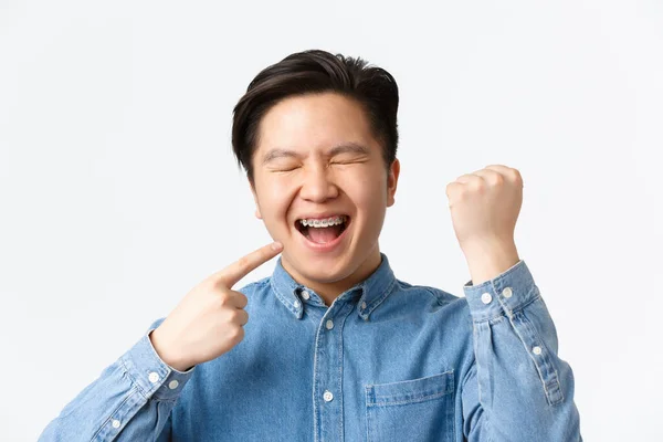 Ortodonti och stomatologi koncept. Närbild av upphetsad och glad asiatisk kille jublar över nya tänder hängslen, pekar på munnen och ler, knytnäve pump, triumferande över vit bakgrund — Stockfoto