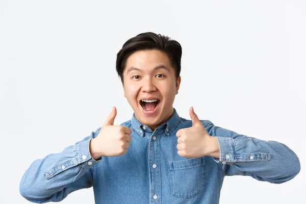 Närbild av framgångsrika vinnande, glad asiatisk man med tänder hängslen ler brett och visar tummen upp i godkännande, prisa stort arbete, säger bra gjort, stående vit bakgrund nöjd — Stockfoto