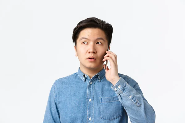 Κοντινό πλάνο του αναποφάσιστου Ασιάτη που απομακρύνεται κατά τη διάρκεια του τηλεφωνήματος, συζητάει και κοιτάζει αλλού μπερδεμένος, στέκεται λευκό φόντο. Ο άνθρωπος κρατώντας smartphone κοντά στο αυτί, παράδοση τροφίμων παραγγελία — Φωτογραφία Αρχείου