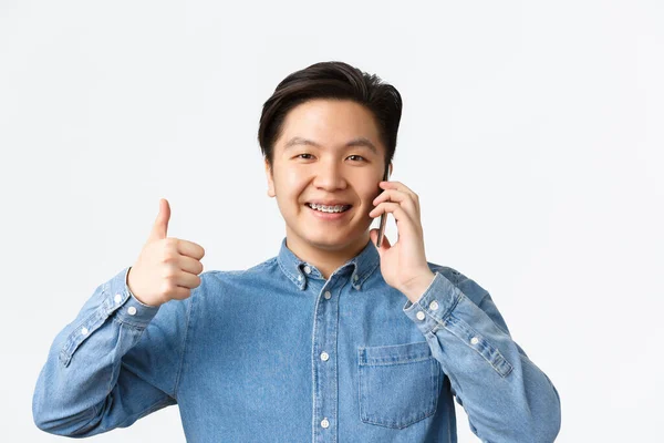 Närbild av glad nöjd asiatisk man visar tummen upp i godkännande när du talar på telefon, få positivt svar eller tillstånd, ler nöjd, rekommendera något, vit bakgrund — Stockfoto