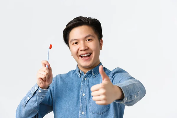 Orthodontics, dental care and hygiene concept. Close-up of satisfied happy asian guy holding toothbrush, smiling with teeth braces and showing thumbs-up in approval, white background — Stock Photo, Image