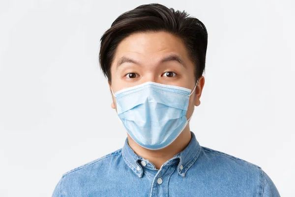 Coronavirus, socialt distans och livsstil koncept. Närbild av upphetsad och fascinerad asiatisk kille bär medicinsk mask och titta nyfiken på kameran, höra intressant förslag, vit bakgrund — Stockfoto