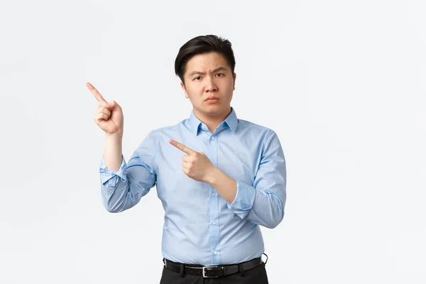 Geschäfts-, Finanz- und Personenkonzept. Enttäuschte Stirnrunzeln asiatischer Geschäftsmann in blauem Hemd zeigt frustriert in die linke obere Ecke, schimpft Mitarbeiter, steht vor weißem Hintergrund unzufrieden — Stockfoto