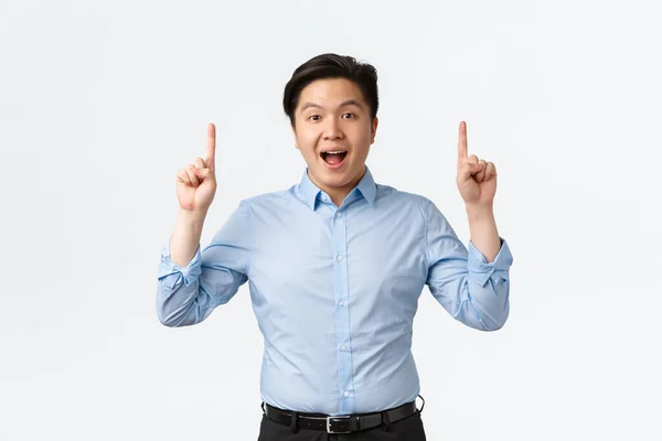Business, finance and people concept. Excited and amazed asian businessman in blue shirt making announcement, pointing fingers up and look camera, telling big news, white background — Stock Photo, Image