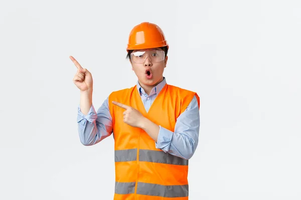 Az építőipar és az ipari dolgozók koncepciója. Meglepett és lenyűgözött ázsiai férfi mérnök, építőipari vezető a gyárban visel biztonsági sisak, fényvisszaverő ruházat, rámutatva a bal felső sarokban — Stock Fotó