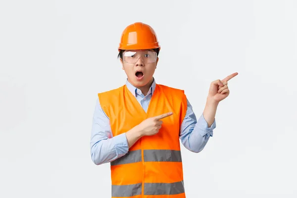 건설 산업과 산업 노동자 개념이다. 공장에서 보호용 투구와 안전유리를 착용하고 일하는 아시아인 남자 건축가, 오른쪽 위를 가리키는 오른쪽 위를 보고 놀라다 — 스톡 사진