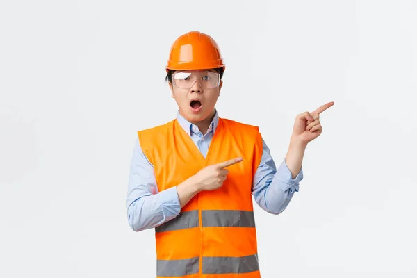 Secteur du bâtiment et concept des travailleurs industriels. Asonished et impressionné asiatique architecte masculin, ingénieur en casque de protection et lunettes de sécurité de travail à l'usine, pointant coin supérieur droit étonné — Photo