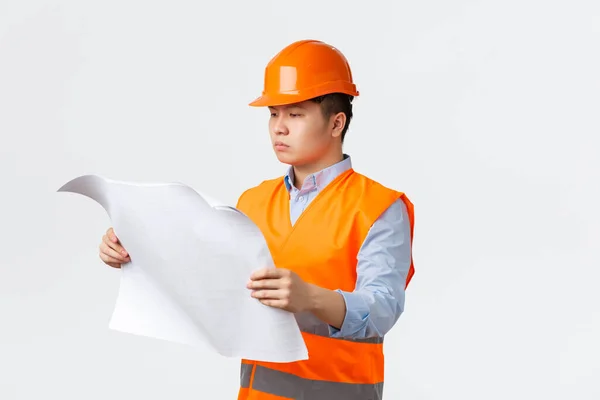 Concepto de sector de la construcción y trabajadores industriales. Gerente de construcción asiática de aspecto serio, arquitecto estudiando planos, mirando el diseño con cara seria, de pie fondo blanco en el casco — Foto de Stock