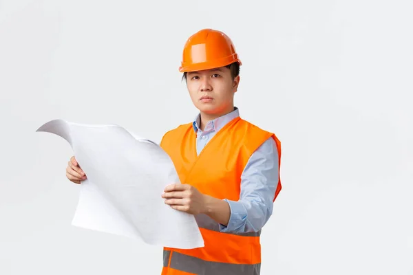 Концепция строительного сектора и промышленных рабочих. Серьёзно выглядящий уверенный азиатский строительный менеджер, инженер в шлеме и светоотражающей куртке, смотрящий на чертеж, проверяющий макет, белый фон — стоковое фото