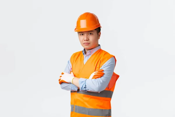 Concepto de sector de la construcción y trabajadores industriales. Confiado ingeniero jefe asiático de aspecto serio, arquitecto en casco y ropa reflectante brazos cruzados y mirar cámara determinada, fondo blanco —  Fotos de Stock