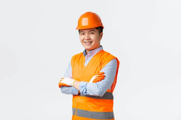 Concepto de sector de la construcción y trabajadores industriales. Confiado joven ingeniero asiático, gerente de construcción en ropa reflectante y casco, brazos cruzados y sonriente descarado, asegurando calidad, fondo blanco —  Fotos de Stock