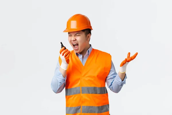 Building sector and industrial workers concept. Angry and pissed-off asian chief engineer scolding workers for mistakes, shouting in walkie-talkie furious, standing in helmet and reflective clothing — Foto Stock