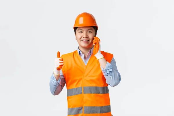 건설 산업과 산업 노동자 개념이다. 건설 책임자는 휴대폰을 통해 수석 엔지니어의 허가나 승인을 받았다. 헬멧을 쓴 아키 텍트는 스마트폰으로 얘기하는 동안 엄지 손가락들을 보여 줍니다. — 스톡 사진