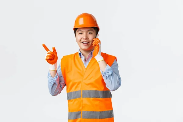 Az építőipar és az ipari dolgozók koncepciója. Mosolygó ázsiai mérnök, építőipari vezető reflektív ruházati és sisak ujjal mutogató balra, miközben beszél a mobiltelefon, fehér háttér — Stock Fotó
