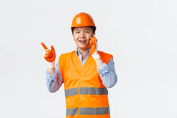 Secteur du bâtiment et concept des travailleurs industriels. Souriant ingénieur asiatique, directeur de la construction dans des vêtements réfléchissants et casque pointant du doigt vers la gauche tout en parlant sur téléphone mobile, fond blanc — Photo