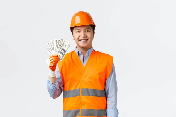 Concepto de sector de la construcción y trabajadores industriales. Feliz sonriente ingeniero asiático, arquitecto en ropa reflectante y casco mostrando dinero con expresión complacida, consiguió el pago de los trabajos de construcción — Foto de Stock