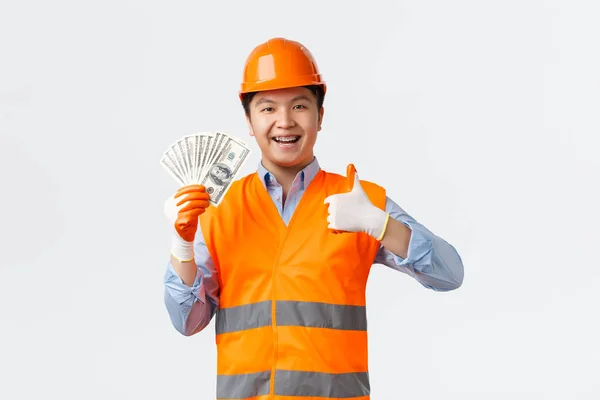 Conceito de sector da construção e trabalhadores industriais. Feliz satisfeito asiático construtor, gerente de construção em capacete e roupas reflexivas mostrando polegares para cima e dinheiro, tem pagamento, fundo branco — Fotografia de Stock