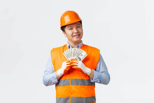Building sector and industrial workers concept. Happy satisfied asian builder, construction manager in reflective clothing and helmet smiling pleased while holding salary, money for work