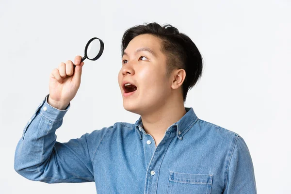 Primer plano del hombre asiático fascinado, investigador en camisa azul, mirando a través de lupa en la esquina superior izquierda con expresión asombrada, encontró algo interesante, fondo blanco — Foto de Stock