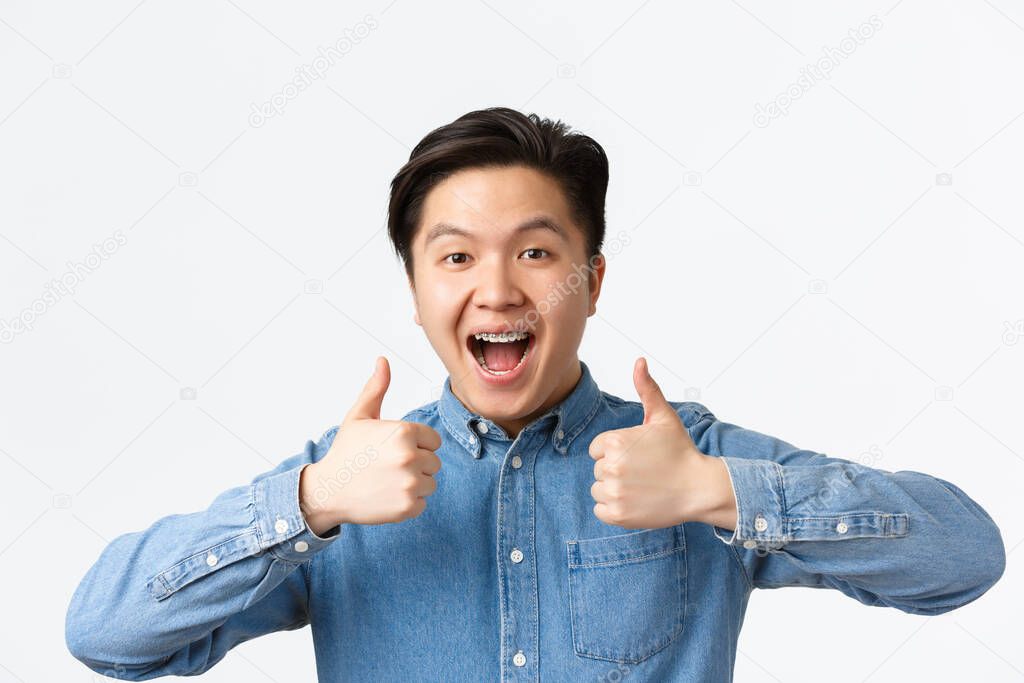 Close-up of successful winning, happy asian man with teeth braces smiling broadly and showing thumbs-up in approval, praising great work, saying well done, standing white background pleased
