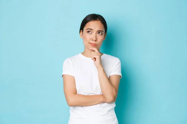 Lifestyle, beauty and shopping concept. Thoughtful and perplexed asian girl thinking, making serious decision, looking upper left corner and pondering, choosing over blue background — Stock Photo, Image