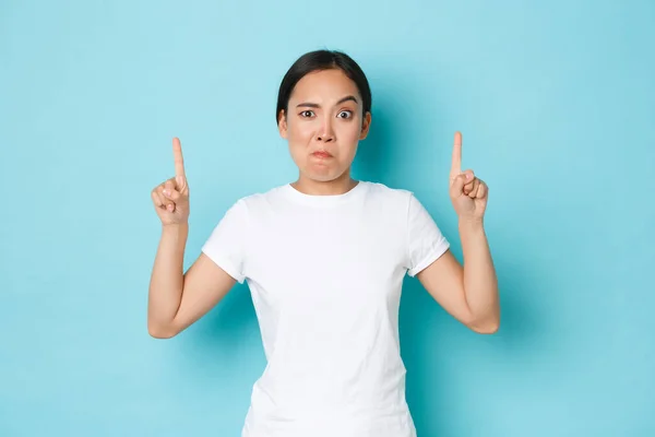 Boos en ontevreden, aziatisch grappig meisje in wit t-shirt, het maken van ingewikkelde, verdachte of gekke grimas, wachtende uitleg, wijzende vingers naar lege ruimte, staan blauwe achtergrond — Stockfoto