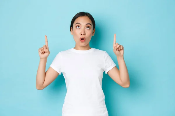 Surprised and impressed asian girl in white t-shirt looking and pointing fingers up at something interesting and amazing. Beautiful korean girl 20s found awesome promo offer, over blue background — Stock Photo, Image