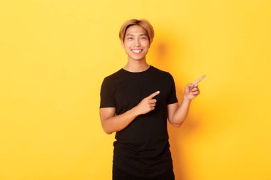 Tatmin olmuş gülümseyen sarışın adam portresi, Asyalı öğrenci parmaklarını sağa gösteriyor, yolu gösteriyor, sarı arka planda duruyor.