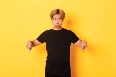 Hüzünlü ve üzgün Asyalı adamın portresi. Pişmanlıkla somurtuyor, pankartı işaret ediyor ve aşağı bakıyor, sarı arka plan.