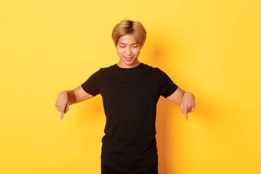 Meraklı, mutlu, sarışın Asyalı bir adamın portresi, mutlu bir gülümsemeyle, sarı arka planda, aşağı bakıyor ve işaret ediyor.
