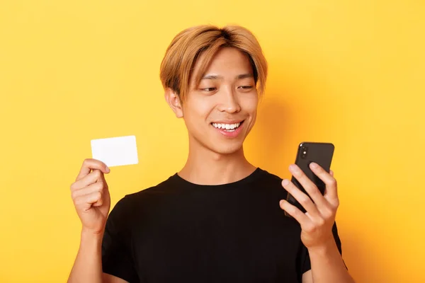 Κοντινό πλάνο του όμορφου κομψού Ασιάτη που ψωνίζει στο διαδίκτυο, κοιτάζει το κινητό τηλέφωνο και χαμογελά, δείχνει πιστωτική κάρτα, στέκεται πάνω από το κίτρινο φόντο — Φωτογραφία Αρχείου