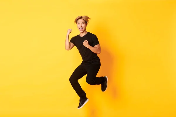 Longitud completa de feliz atractivo asiático chico en negro ropa saltar y celebrar la victoria, lograr la meta, de pie fondo amarillo, triunfante — Foto de Stock