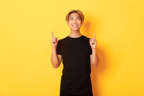 Snygg leende asiatisk man i svart t-shirt pekar fingrarna upp, tittar på banner nöjd, gul bakgrund — Stockfoto