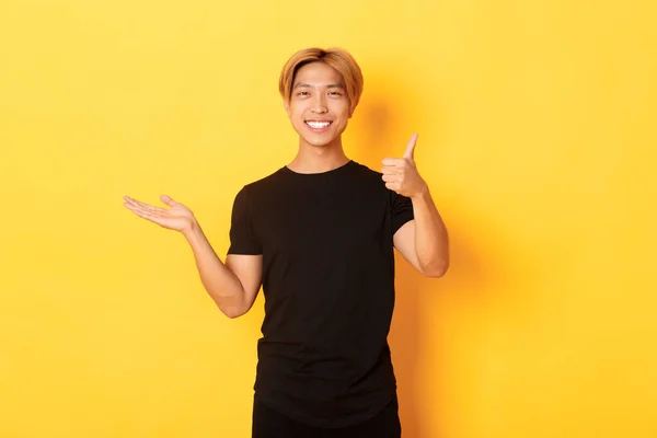 Spokojenej a šťastnej atraktivní korejskej chlápek, co se usmívá, radostně ukazuje palce nahoru, drží něco po ruce, stojí na žlutým pozadí — Stock fotografie