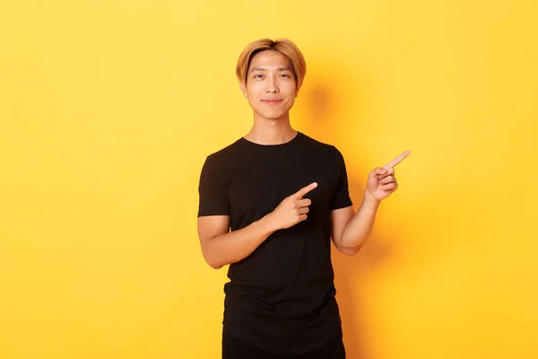 Usmívající se asijský student s blond vlasy, ukazující prsty vpravo, ukazující logo, stojící žluté pozadí — Stock fotografie