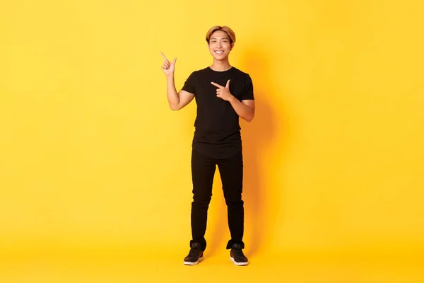Full-length di sorriso attraente coreano ragazzo puntando il dito verso sinistra, mostrando logo, sfondo giallo — Foto Stock