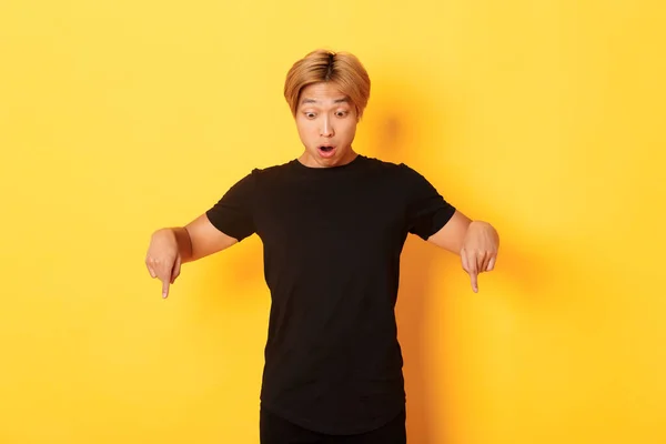 Portrait of surpirsed asian guy gasping amazed, pointing fingers and looking down at banner, standing yellow background — Stock Photo, Image