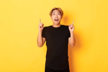 Etkileyici Asyalı sarışın adam, siyah tişört giyiyor, heyecanlı ve işaret ediyor, sarı arka plan