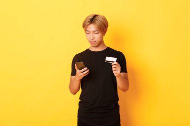 Siyah tişörtlü genç yakışıklı Asyalı adam internetten alışveriş yapıyor, kredi kartı tutuyor ve akıllı telefon kullanıyor, sarı arka planda duruyor.