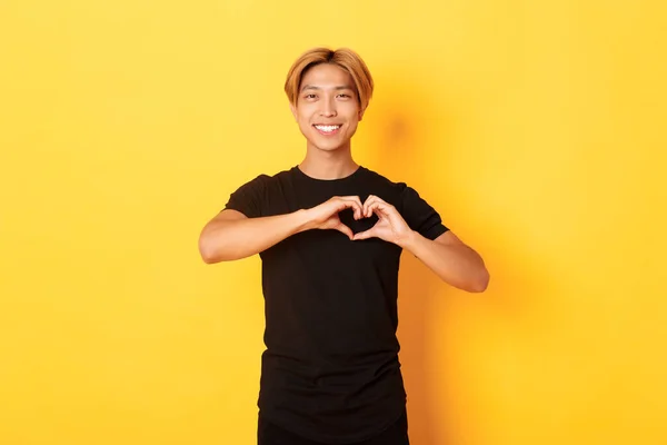 Portrét půvabného pohledného blonďatého Asiata, ukazující gesto srdce a úsměv, stojící na žlutém pozadí — Stock fotografie