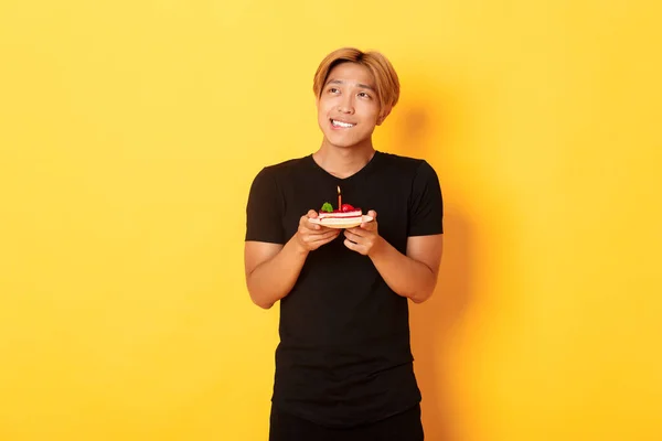 Retrato de bonito sonhador asiático cara olhando canto superior esquerdo e pensando, fazendo desejo ao celebrar aniversário e segurando b-dia bolo, fundo amarelo — Fotografia de Stock