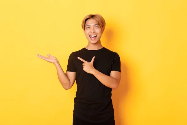 Portrét pohledného usměvavého Asiata držícího něco na ruce a ukazujícího na to prstem, stojící žluté pozadí potěšené — Stock fotografie