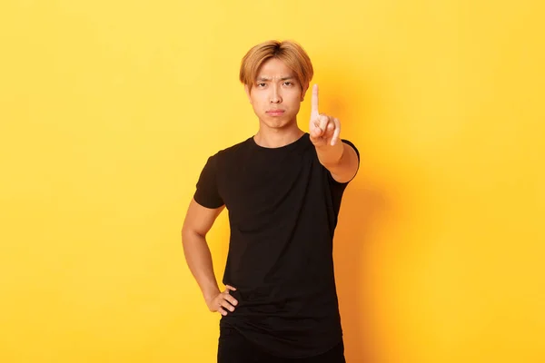 Portrait of serious-looking disappointed asian man shaking finger to scold someone, standing yellow background