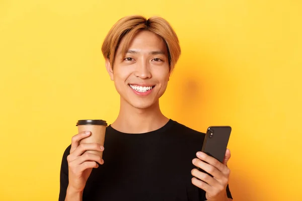 Primer plano de guapo joven asiático sonriendo feliz, usando teléfono inteligente y beber café para llevar, de pie sobre fondo amarillo — Foto de Stock