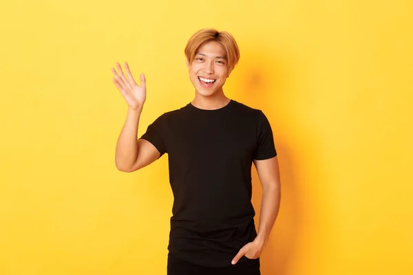 Retrato de bonito amigável asiático cara em roupa preta, acenando mão para dizer Olá e sorrindo, cumprimentando alguém, de pé fundo amarelo — Fotografia de Stock