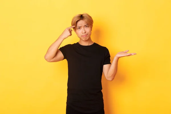 Decepcionado chico asiático con pelo rubio, levantando la mano desconcertado y regañando a alguien actuando estúpido, de pie fondo amarillo — Foto de Stock