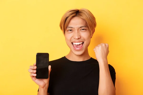 Close-up de feliz regozijo asiático cara mostrando tela do smartphone e dizendo sim, punho bomba como triunfar, ganhar ou alcançar objetivo, fundo amarelo — Fotografia de Stock