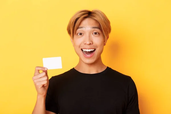Közelkép izgatott meglepett jóképű ázsiai srác, mosolygós csodálkozott, és mutatja a hitelkártya, álló sárga háttér — Stock Fotó