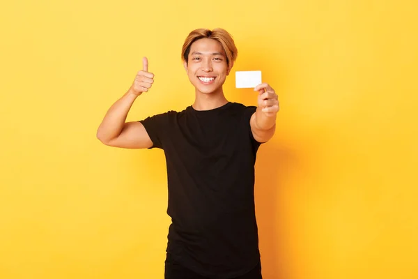 Spokojenost pohledný asijský chlap usmívá potěšen a ukazuje kreditní kartu, palce nahoru v souhlasu, stojící žluté pozadí — Stock fotografie