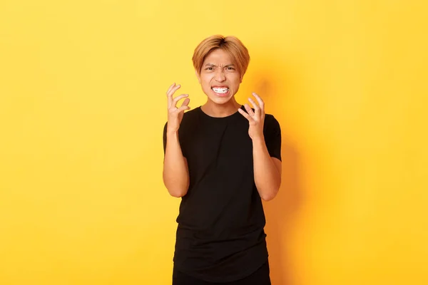 Portrait of angry and pissed-off asian blond guy, clenching hands furious and grimacing, standing over yellow background — Stock Fotó